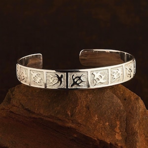 May include: A silver cuff bracelet with a series of square panels, each featuring a unique tribal design. The bracelet is open-ended and rests on a brown, textured surface. The metal has a polished, reflective finish.