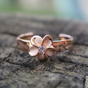 Puede incluir: Un anillo de dedo del pie de oro rosa con un diseño de flor blanca y un pequeño strass transparente en el centro.