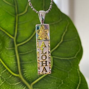 May include: A silver chain necklace with a gold pendant. The pendant is a rectangular shape with the word "ALOHA" engraved in gold letters. The pendant also features a gold flower design.