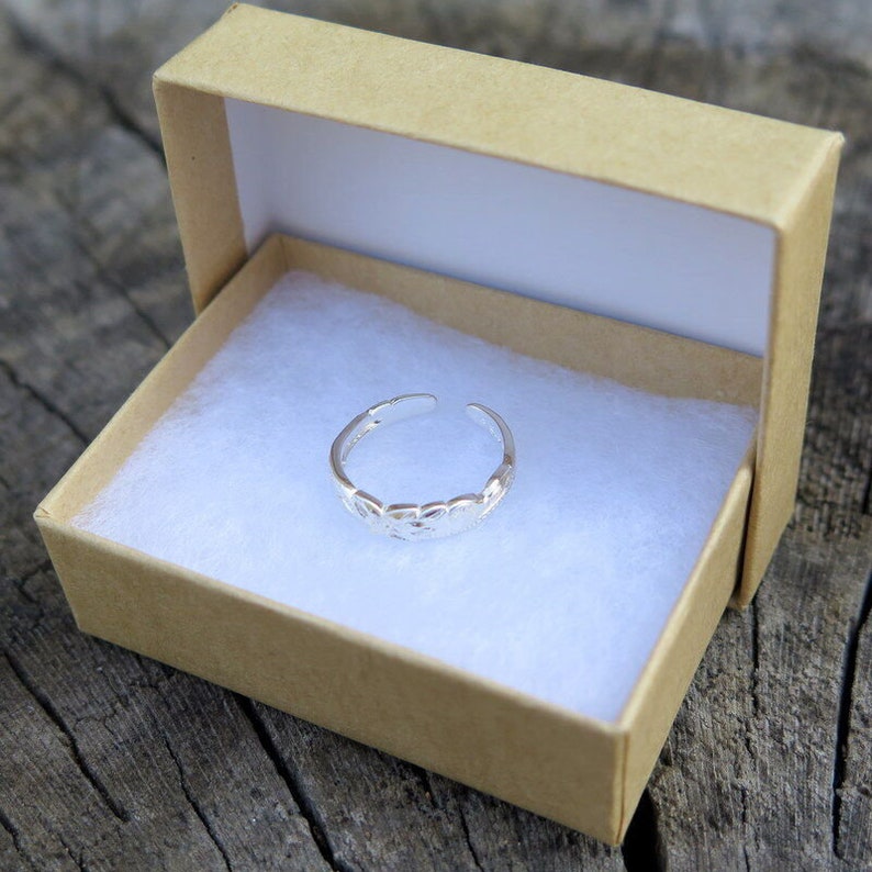 May include: A silver toe ring with a delicate, patterned design sits in a brown box with white lining.