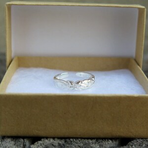 May include: A silver toe ring with an intricate floral design. The ring is displayed in a small, brown gift box.