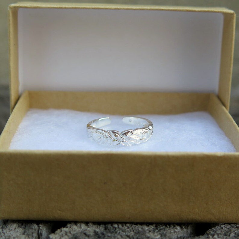 May include: A silver toe ring with an intricate floral design. The ring is displayed in a small, brown gift box.