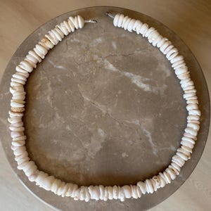 Könnte beinhalten: Eine Halskette aus weißen und hellrosa Muschelstücken. Die Muscheln sind zu einer kreisförmigen Form aufgereiht. Die Halskette liegt auf einer braunen Marmoroberfläche.