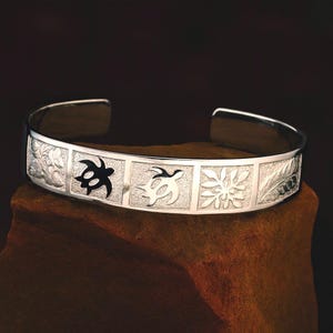 Puede incluir: Una pulsera de plata con paneles grabados. Los paneles presentan diseños florales y de tortugas. La pulsera se muestra sobre una superficie marrón sobre un fondo oscuro. La pulsera está abierta en los extremos.