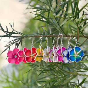 Puede incluir: Una colección de pendientes con forma de flor en colores del arcoíris, incluyendo rosa, rojo, amarillo, blanco y azul, cada uno con un cristal central. Los pendientes cuelgan de ganchos plateados y se exhiben en una rama frondosa.