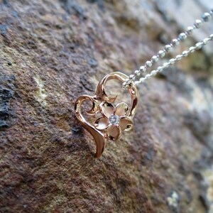 Puede incluir: Un collar con un colgante en forma de coraz&oacute;n de oro rosa, adornado con un delicado dise&ntilde;o floral y una sola piedra transparente en el centro.