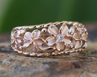 Puede incluir: Un anillo de oro rosa con un delicado diseño floral con cinco flores de plumeria. El anillo está engastado con un solo diamante pequeño en el centro del anillo.
