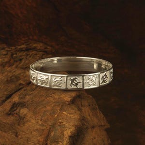 Puede incluir: Un brazalete plateado con un patrón repetitivo de símbolos estilizados. El brazalete se muestra sobre un trozo de madera marrón oscuro. Los símbolos están grabados en secciones rectangulares.