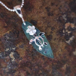 Puede incluir: Un collar de plata con un colgante de tabla de surf verde y blanco con una tortuga plateada y una flor blanca.