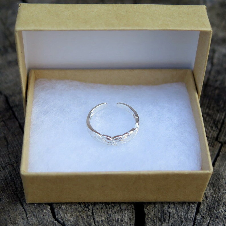 May include: A silver toe ring with a delicate floral design, displayed in a brown box with white lining.