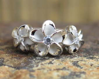Può includere: Un anello d'argento con tre fiori di frangipane bianchi, ognuno con una piccola pietra preziosa trasparente al centro.