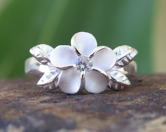 Può includere: Un anello d'argento con un designo a forma di fiore di frangipane bianco e un cristallo trasparente al centro.
