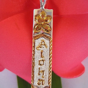May include: A silver pendant necklace with a gold flower design at the top. The pendant has the word "Aloha" in gold lettering. The pendant is rectangular with a textured silver center and gold rope-like edges.