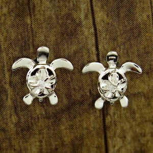 May include: A pair of silver earrings featuring a detailed turtle design with a flower in the center, accented with a clear crystal.