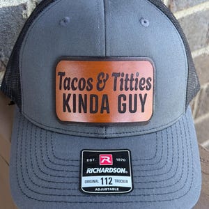 May include: Gray trucker hat with a black mesh back. The hat features a brown leather patch with the text "Tacos & Titties Kinda Guy". The Richardson brand label is visible.