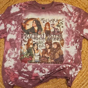 May include: A purple tie-dye t-shirt with a graphic of four women in witch costumes. The text "Practical Magic" is printed on the shirt, along with the quote "There's a little bit of witch in all of us."