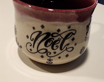 May include: Cream ceramic mug with a burgundy rim and handle. The mug features the word "Noël" in elegant black script, surrounded by small floral designs. A decorative holiday-themed mug.