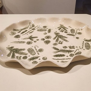 Puede incluir: Un plato de servir de cerámica blanca con un borde festoneado. El plato está decorado con un patrón verde y blanco de pinos, piñas y bastones de caramelo.