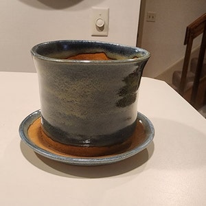 Puede incluir: Una maceta de cerámica azul y marrón con un platillo a juego. La maceta tiene un acabado esmaltado con un patrón moteado.