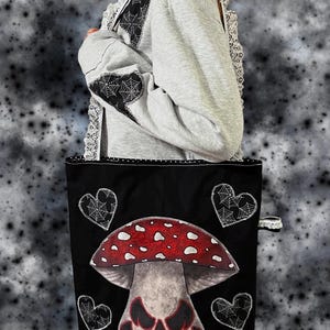 May include: Black tote bag with a red and white mushroom design and a jack-o'-lantern face. The bag has black heart accents with spiderwebs. A grey hoodie with black spiderweb accents and white lace trim is visible.