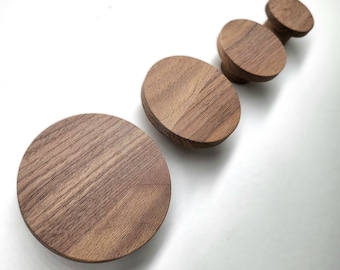 May include: A collection of four wooden cabinet knobs in varying sizes, arranged diagonally on a white surface. The knobs are a warm brown color, showcasing the natural wood grain. The largest knob is a flat disc, while the others have a rounded shape.