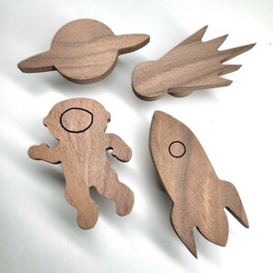 May include: Set of five wooden wall hooks shaped like a rocket, astronaut, comet, and two planets. The hooks are made of walnut wood and have a natural finish.