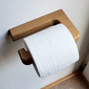 May include: A wooden toilet paper holder mounted on a white wall. The holder is made of light-colored wood and has a simple, modern design. A roll of white toilet paper is in place, ready for use.