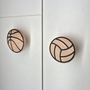 Könnte beinhalten: Zwei Holzschrankknöpfe, einer als Basketball und der andere als Volleyball gestaltet. Die Knöpfe sind aus hellem Holz mit schwarzen Umrissen gefertigt und an einer weißen Schranktür befestigt.