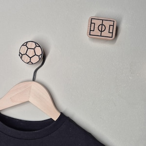 May include: Wooden wall hooks with a soccer ball and a soccer field design. The soccer ball hook is round with black pentagon shapes. The soccer field hook is rectangular with a black field outline. A black shirt hangs on a wooden hanger.