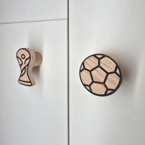 以下が含まれることがあります： 木製のキャビネットノブが2つ。1つは黒いアウトラインのトロフィーの形です。もう1つは黒いアウトラインのサッカーボールのデザインです。どちらも白いキャビネットドアに取り付けられています。