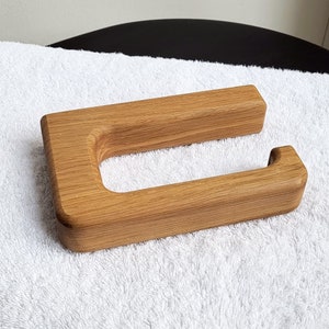 May include: A wooden toilet paper holder with a natural wood grain finish. The holder has a rectangular shape with a curved opening for the toilet paper roll. The wood is light brown in color and appears to be smooth to the touch.