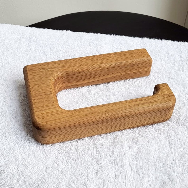 May include: A wooden toilet paper holder with a natural wood grain finish. The holder has a rectangular shape with a curved opening for the toilet paper roll. The wood is light brown in color and appears to be smooth to the touch.