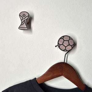 Könnte beinhalten: Zwei Wandhaken aus Holz. Einer ist in Form einer Trophäe mit schwarzen Umrissen. Der andere ist ein Fußball-Design mit schwarzen Umrissen. Unter dem Fußballhaken hängt ein Holzkleiderbügel mit einem schwarzen T-Shirt.