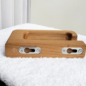 May include: A rectangular wooden phone holder with a carved groove and two metal keyhole brackets for mounting. The holder is made of light brown wood and has a natural wood grain pattern. The brackets are secured with silver screws.