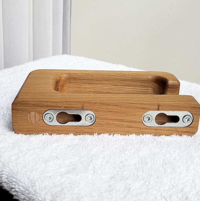 May include: A rectangular wooden phone holder with a carved groove and two metal keyhole brackets for mounting. The holder is made of light brown wood and has a natural wood grain pattern. The brackets are secured with silver screws.