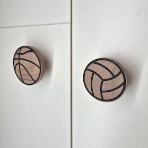 May include: Two wooden cabinet knobs, one designed as a basketball and the other as a volleyball. The knobs are light brown with black outlines, mounted on a white cabinet door. The basketball knob is on the left, and the volleyball knob is on the right.