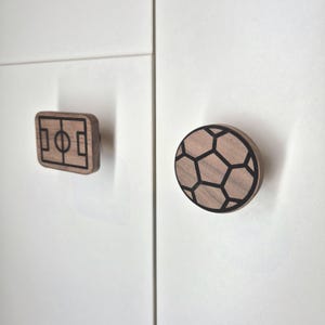 May include: Two wooden cabinet knobs. One is rectangular with a football pitch design, the other is round with a football design. Both knobs have a light brown wood colour with black outlines, mounted on a white cabinet.
