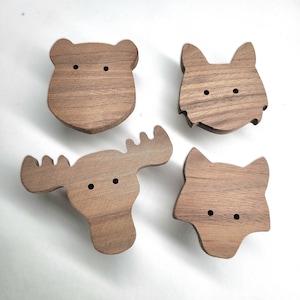May include: Four wooden animal-shaped cabinet knobs. The set includes a bear, a wolf, a moose, and a fox. Each knob is carved from a light brown wood and features two small, round holes for eyes. These knobs are ideal for adding a touch of whimsy to furniture.