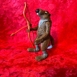May include: A brown anthropomorphic rat action figure standing upright, featuring a textured fur pattern, a grey chest patch, and grey wraps around its feet. The figure holds a brown plastic bow in its right hand, with a black nose and white teeth.