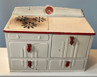 May include: A vintage white toy stove with red accents. The stove features a cooktop with three burner designs, four drawers with red handles, and a red-trimmed top. The stove has an off-white finish with some wear.