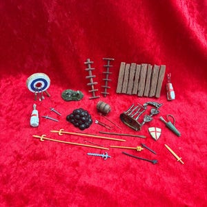 May include: A collection of miniature toy accessories, including a target, ladders, a barrel, arrows, spears, a shield, and a sword. The items are in various colors, such as brown, blue, and yellow, and are arranged on a red surface.