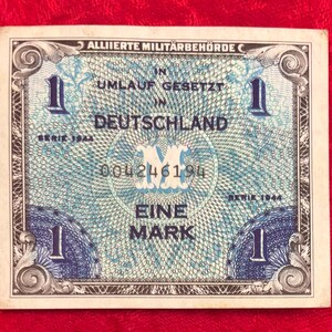 May include: A vintage German banknote, featuring the text "ALLIIERTE MILITÄRBEHÖRDE" and "EINE MARK". The note has a blue and white design with the number "1" in each corner. The serial number "004246194" is visible.