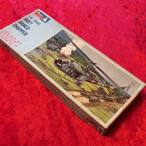 May include: A model kit box for a 1/4 scale Huey Armed Chopper. The box features a colorful illustration of the helicopter in flight, with the words "Huey Armed Chopper" and "Army" printed on the box.