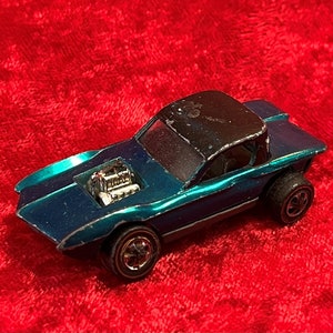 May include: A teal blue Hot Wheels toy car with a black roof and exposed engine. The car has black wheels and a silver chassis.