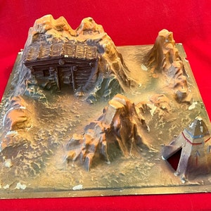May include: A vintage diorama depicting a Western landscape with a log cabin, rock formations, and a teepee. The base is a square with a painted, textured surface. The cabin and teepee are brown and tan, with the mountains in shades of brown and beige.