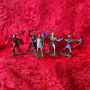 May include: Five toy knight figurines in various poses, each with a weapon. The knights are painted in shades of silver, blue, red, and gold, and stand on green bases. One knight holds a bow and arrow, while others wield swords and spears.