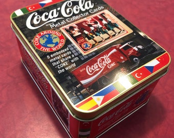 May include: A rectangular Coca-Cola metal collector's card tin. The tin is red with gold trim and features the Coca-Cola logo and various graphics, including a Coca-Cola truck and flags. Text on the tin reads "Coke Around the World."