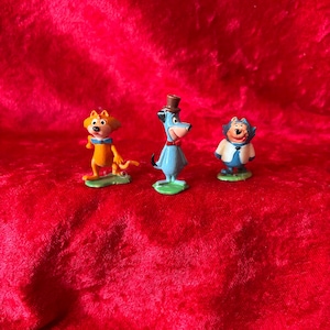 May include: Three small cartoon character figurines standing on green bases. The left figure is an orange cat with a blue bowtie. The center figure is a blue dog wearing a brown top hat and red bowtie. The right figure is a blue and white creature.