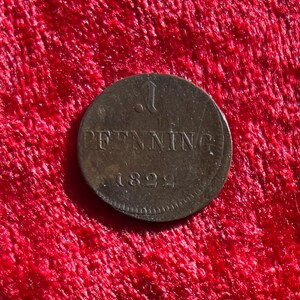 May include: A close-up of a bronze coin with the text "1 PENNING" and the year "1822". The coin is circular and has a slightly worn appearance, set against a red background.