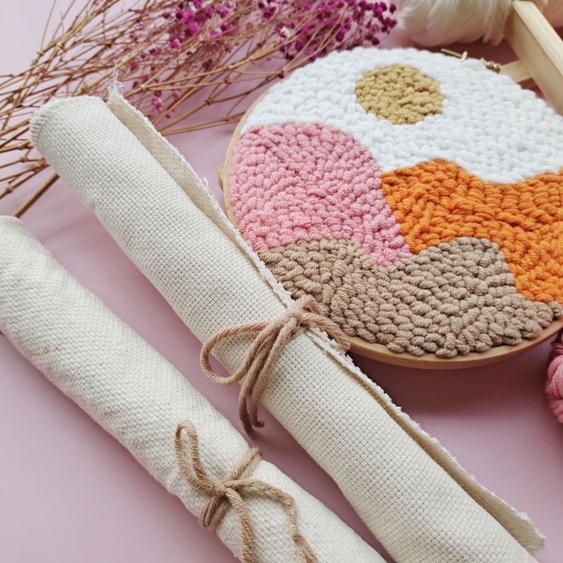 May include: Two rolls of white canvas fabric, tied with twine, are displayed on a pink surface. A completed punch needle embroidery hoop with a geometric design in pink, orange, brown, and white is in the background.